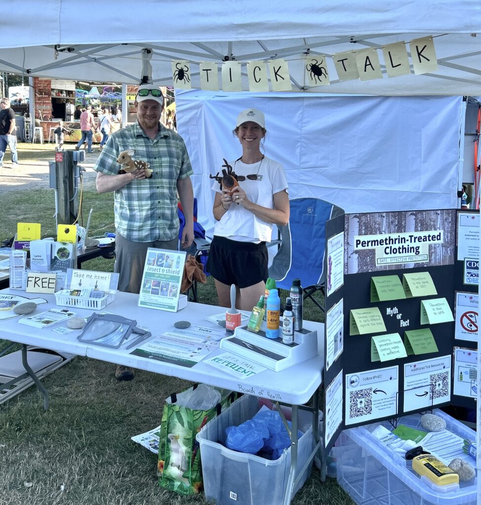 Patrick and Lea smiling behind a table of outreach materials on all things ticks.